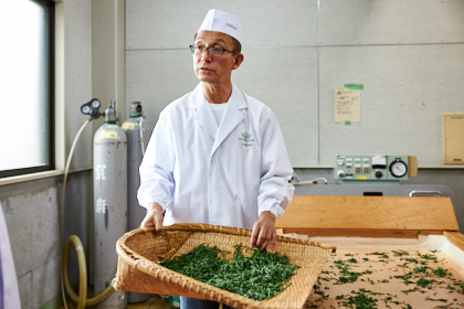 茶葉入りの竹ざるを持つ白衣姿の男性